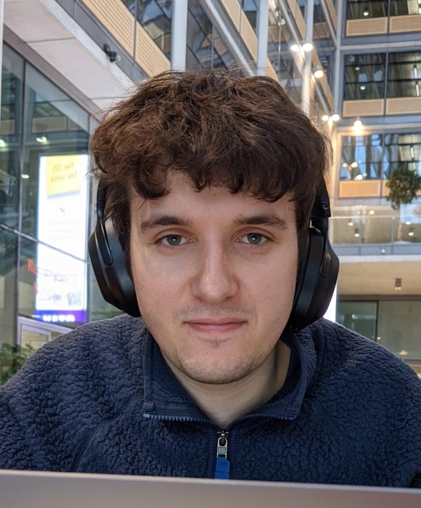 Jack wearing a blue jumper and black headphones, peering over his laptop at the camera